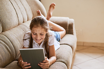 Buy stock photo Shot of a cheerful young girl browsing on a digital tablet while lying on a sofa at home during the day