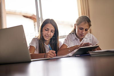 Buy stock photo Shot of two cheerful young girls doing homework together around a table while making use of a digital tablet inside at home during the day