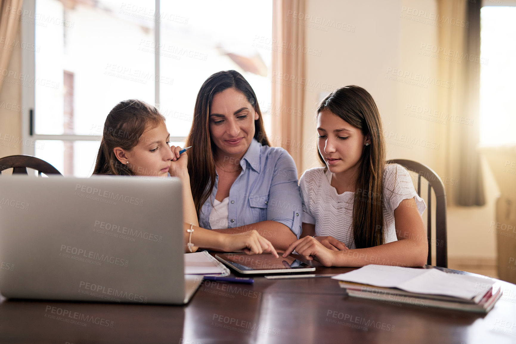Buy stock photo Shot of two cheerful young girls doing homework around a table while getting help from their mother at home during the day
