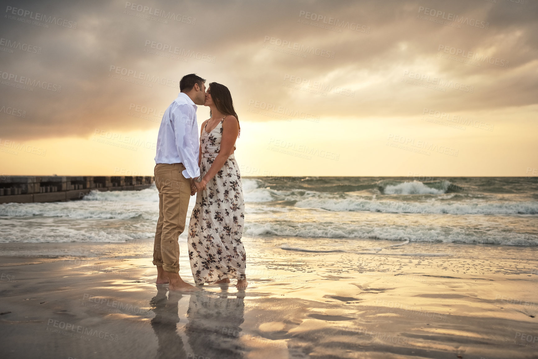 Buy stock photo Full length shot of an affectionate young couple kissing while standing on the beach