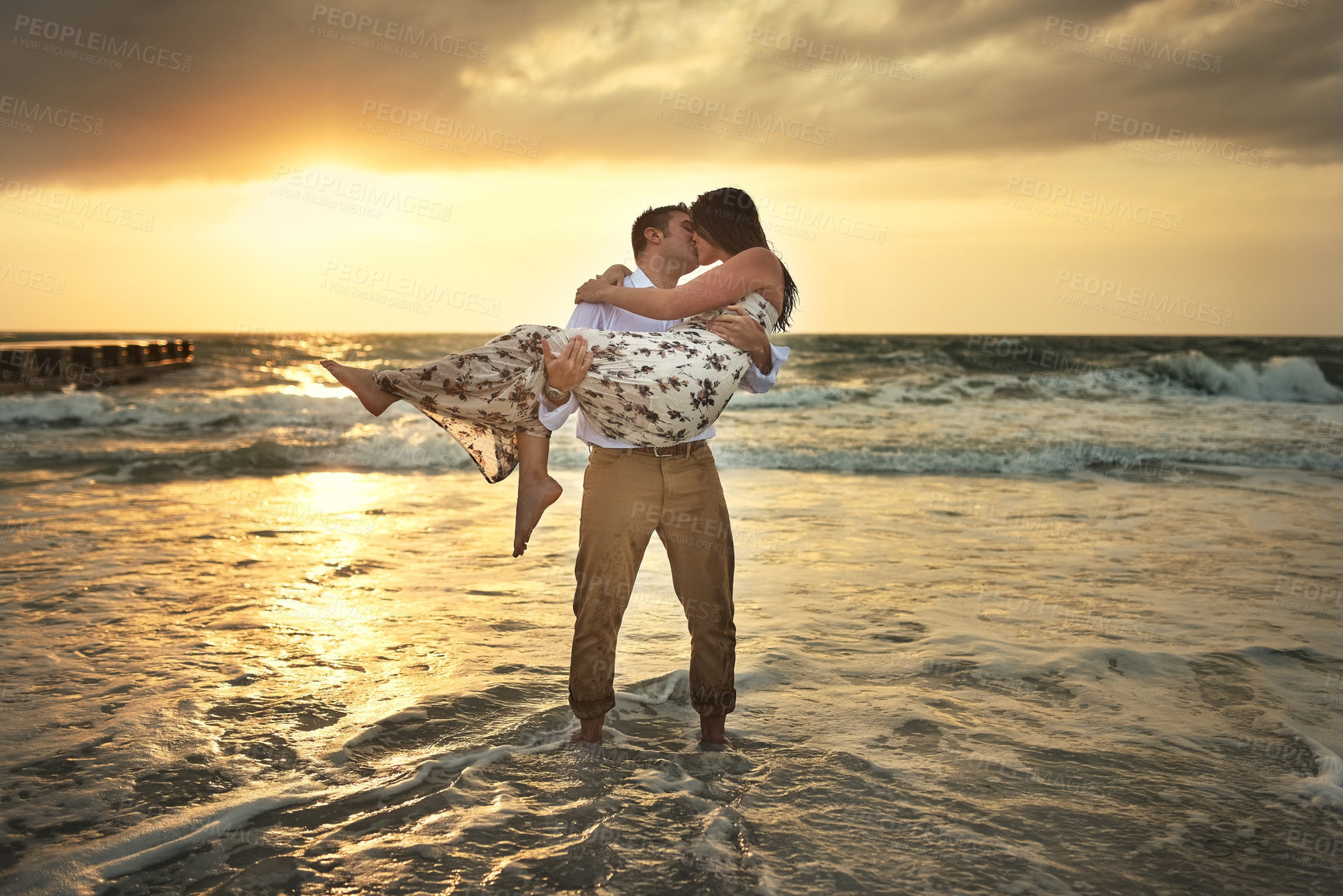 Buy stock photo Couple, kiss and sunset on beach sand, holding wife and together on vacation for connection. People, ocean waves and carrying woman for support in nature, peace and honeymoon date on tropical island