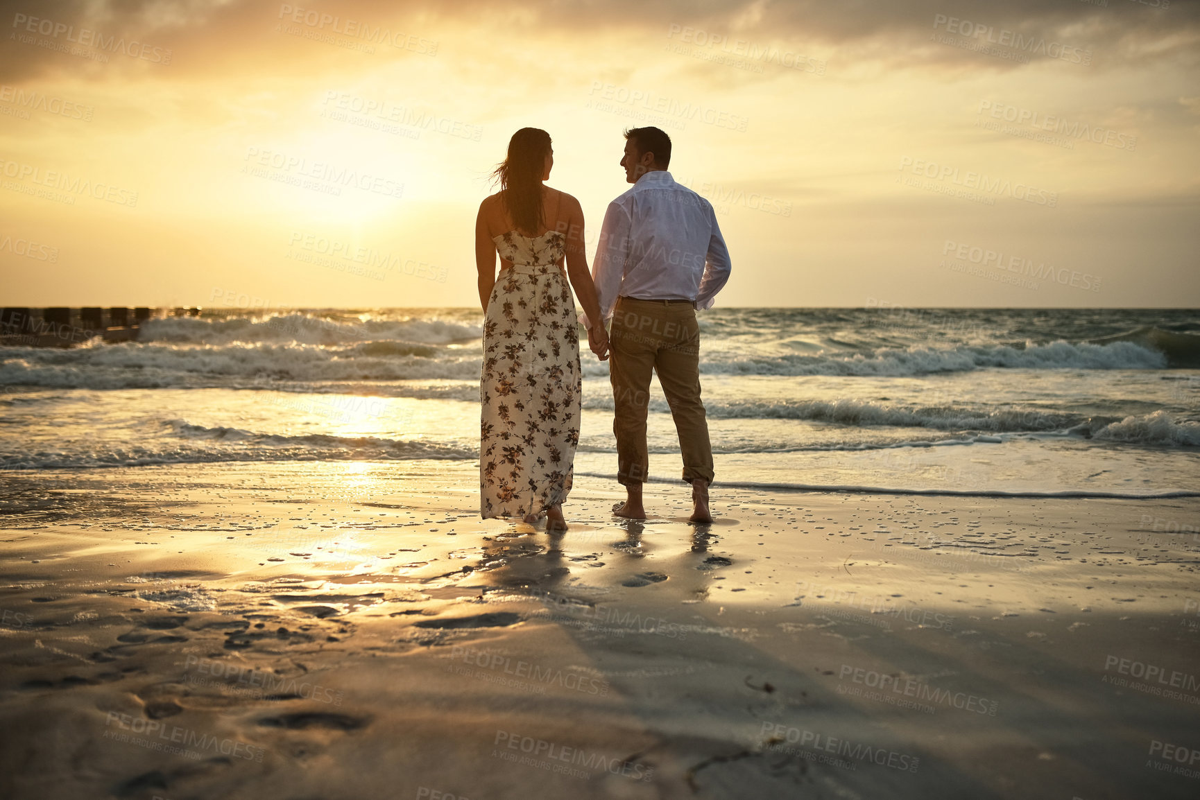 Buy stock photo Rearview shot of an affectionate young couple holding hands while standing face to face on the beach