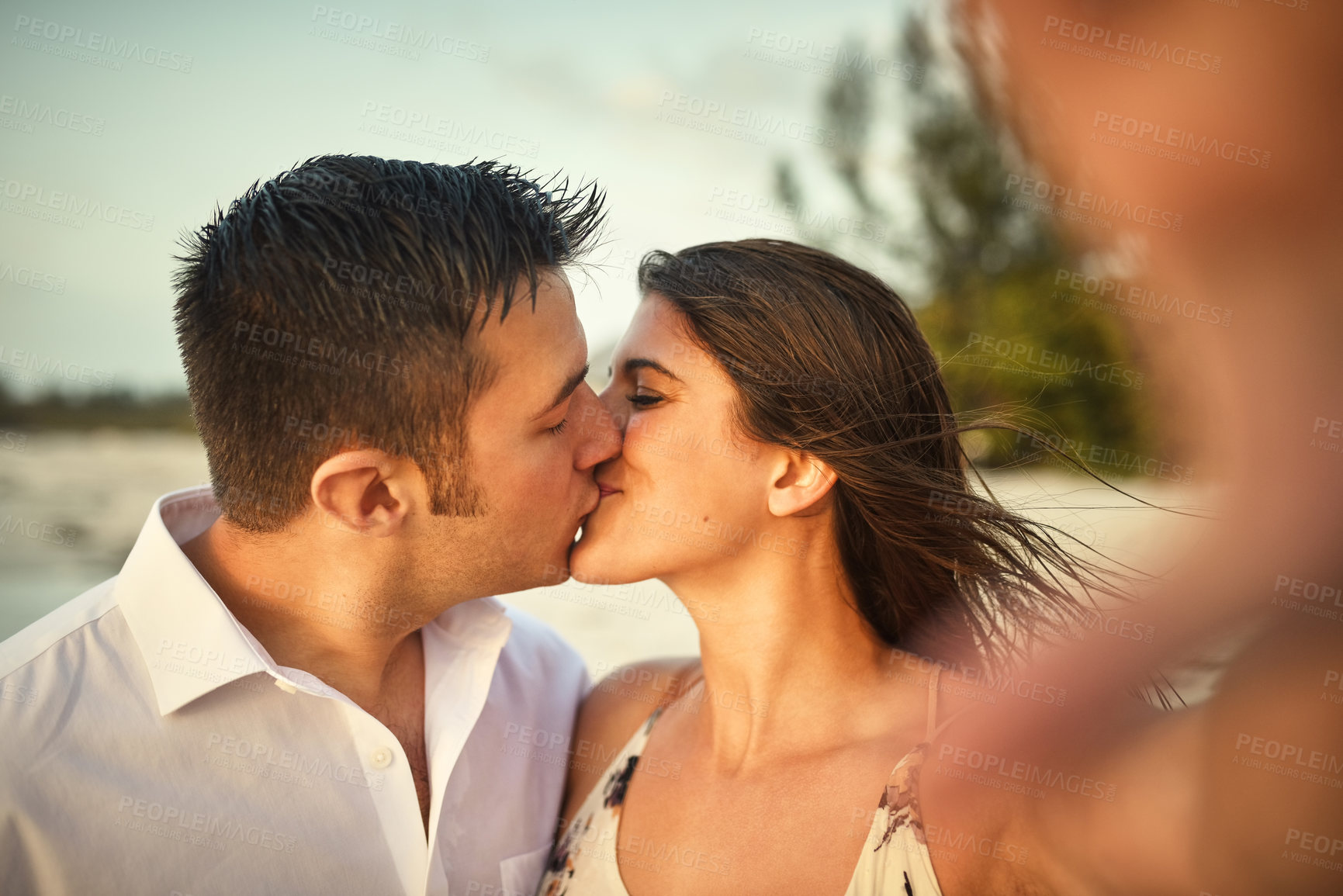 Buy stock photo Cropped shot of an affectionate young couple kissing while taking selfies on the beach