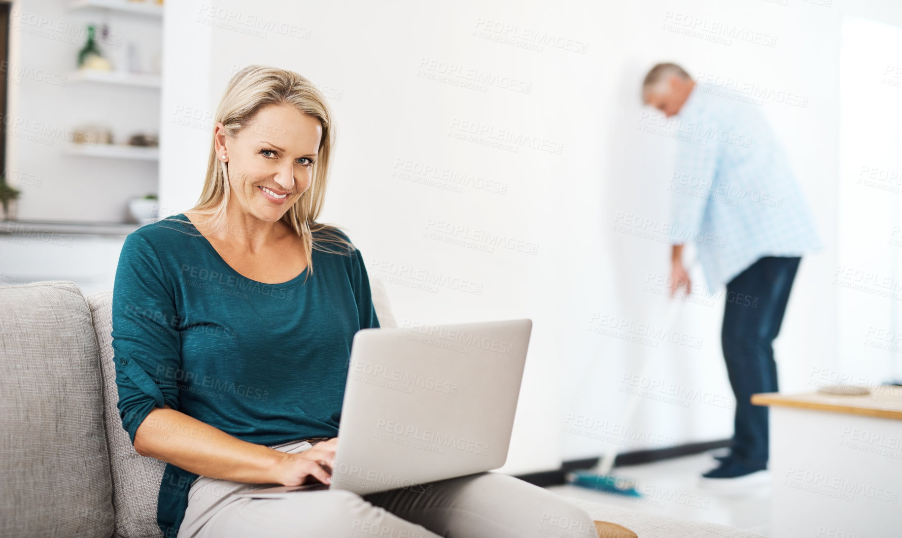Buy stock photo Shot of a mature woman using a laptop at home while her husband sweeps in the background