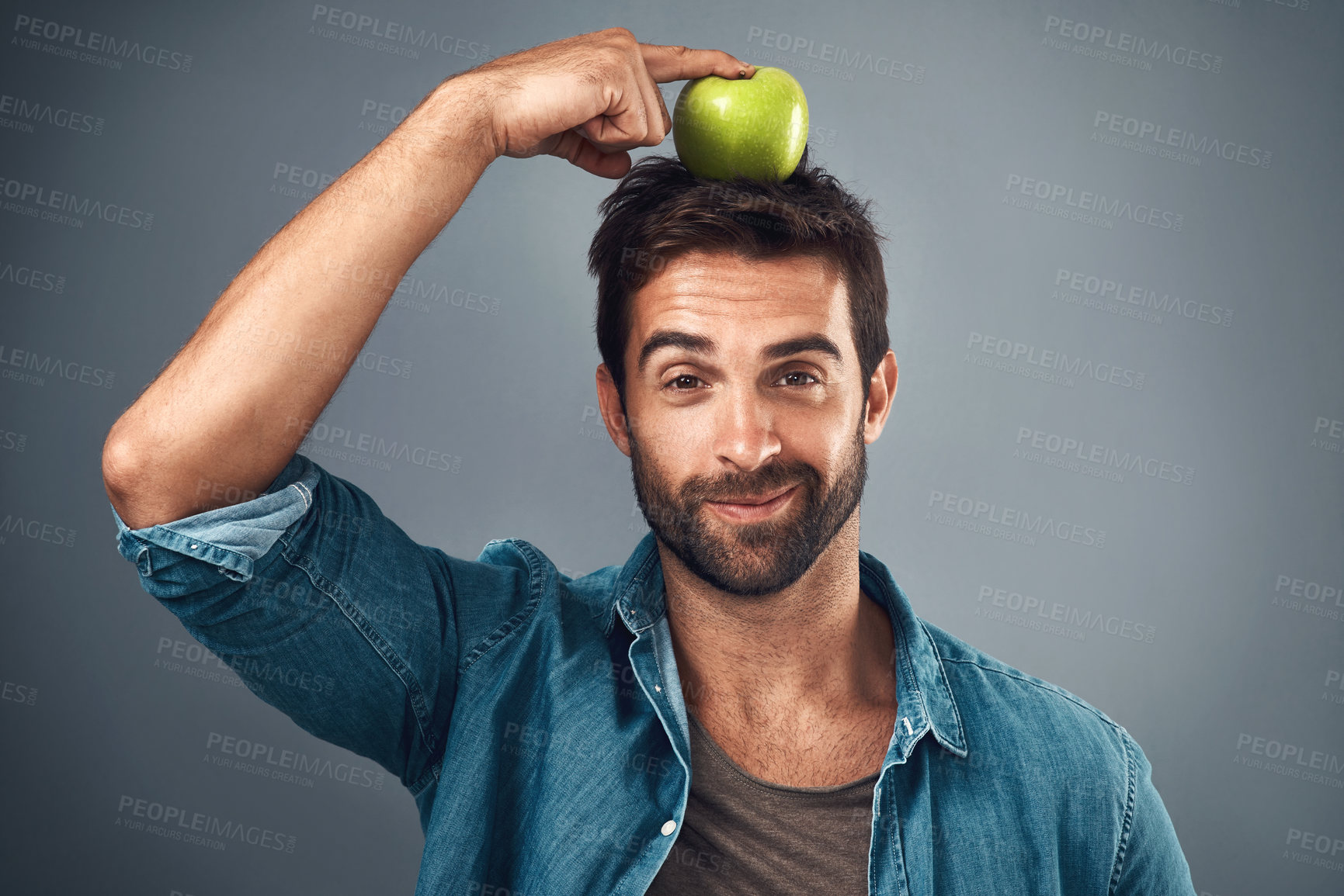 Buy stock photo Studio shot of a handsome young man balancing an apple on his head against a grey background