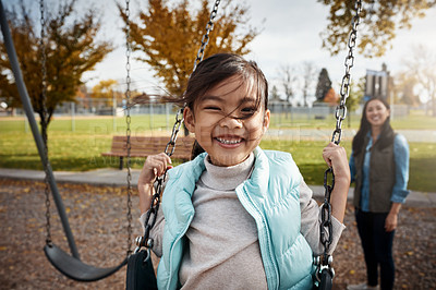 Buy stock photo Portrait of a little girl playing on a swing at the park with her mother in the background