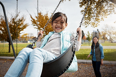 Buy stock photo Portrait of a little girl playing on a swing at the park with her mother in the background