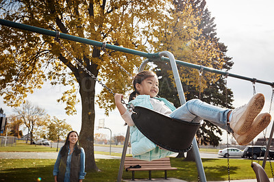 Buy stock photo Shot of a little girl playing on a swing at the park with her mother in the background