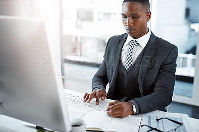 Buy stock photo Shot of a young businessman using a computer and writing notes at his desk in a modern office