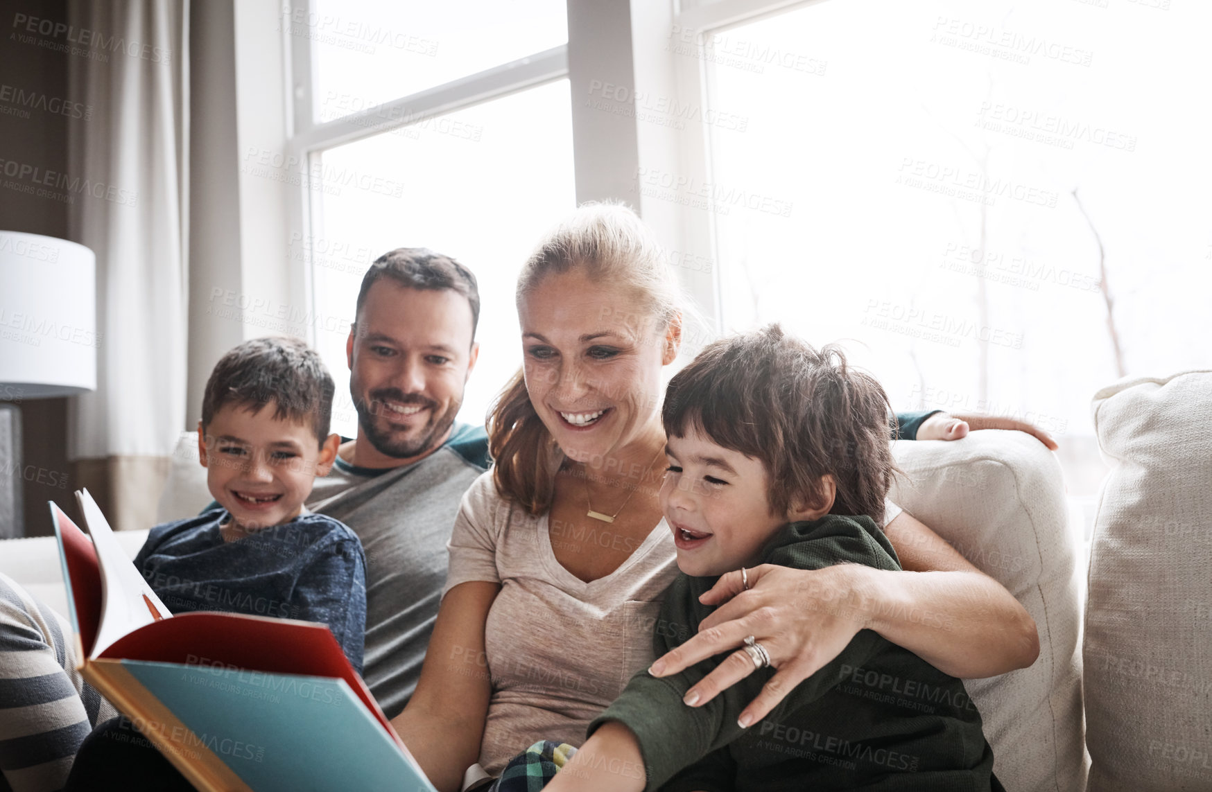 Buy stock photo Shot of two adorable brothers reading a book together with their parents while relaxing on the sofa at home