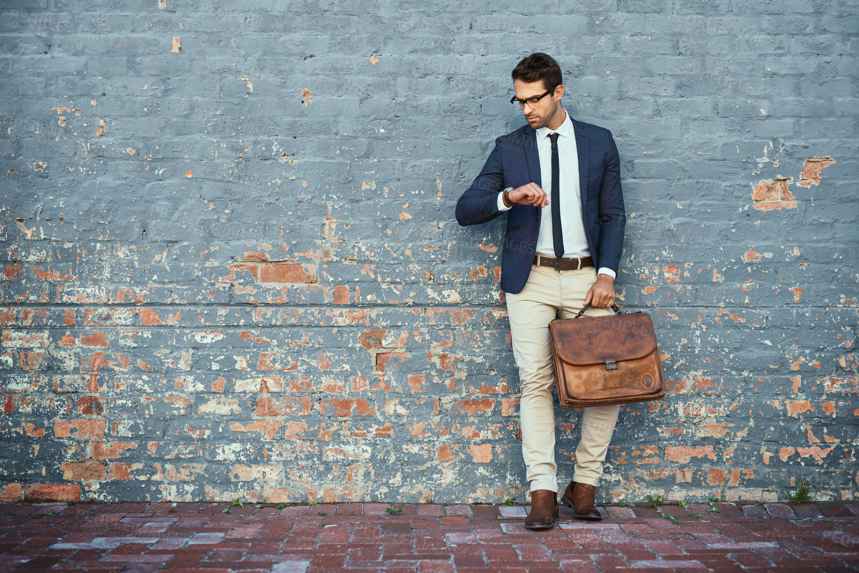 Buy stock photo Full length shot of a handsome young businessman checking the time while standing against a grey facebrick wall