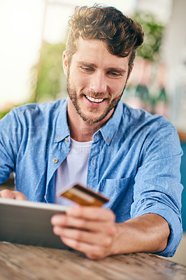 Buy stock photo Shot of a cheerful young man doing online shopping on his digital tablet and holding his credit card while being seated at a table