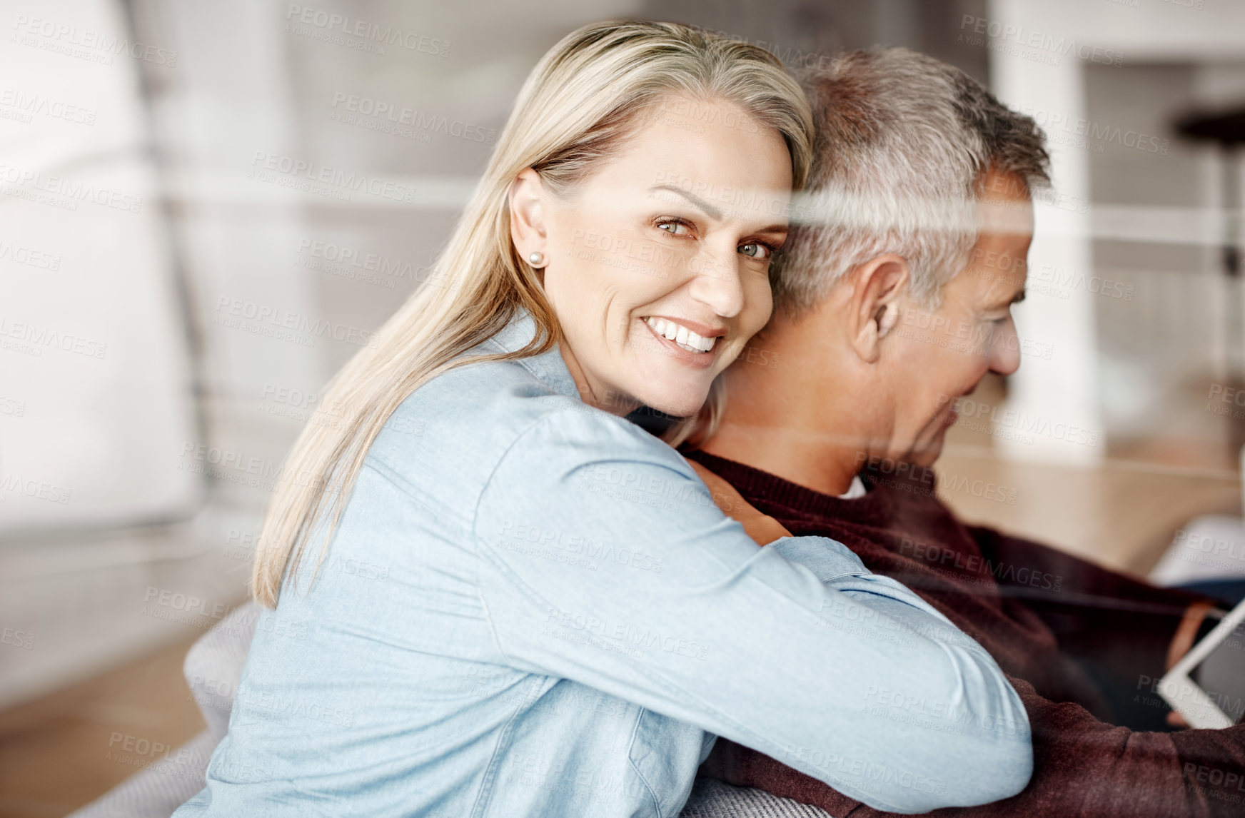 Buy stock photo Shot of a mature woman hugging her husband while he uses a digital tablet on the sofa at home