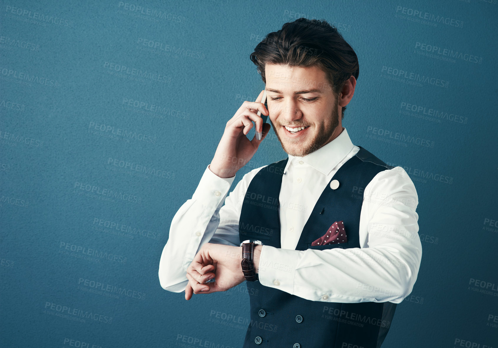 Buy stock photo Studio shot of a handsome young businessman checking the time while making a phonecall against a blue background