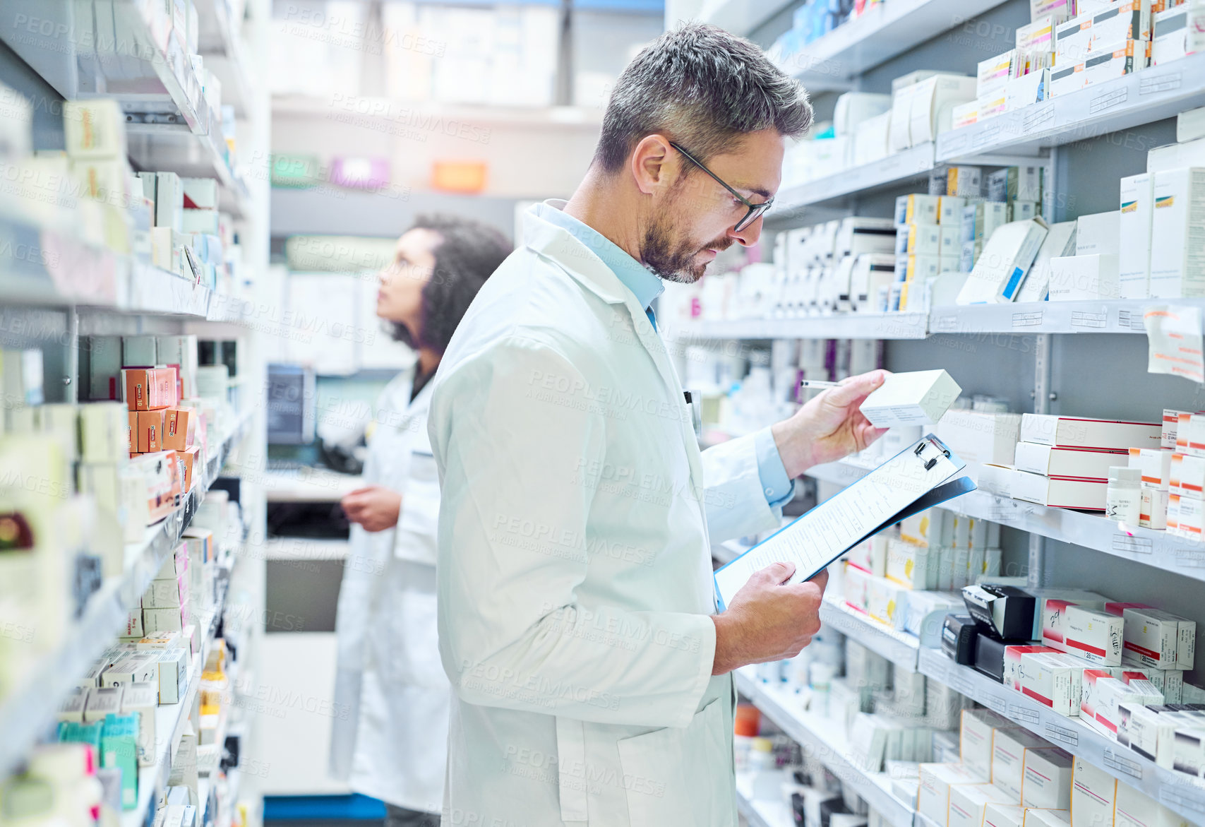 Buy stock photo Shot of a mature man doing inventory in a pharmacy with his colleague in the background