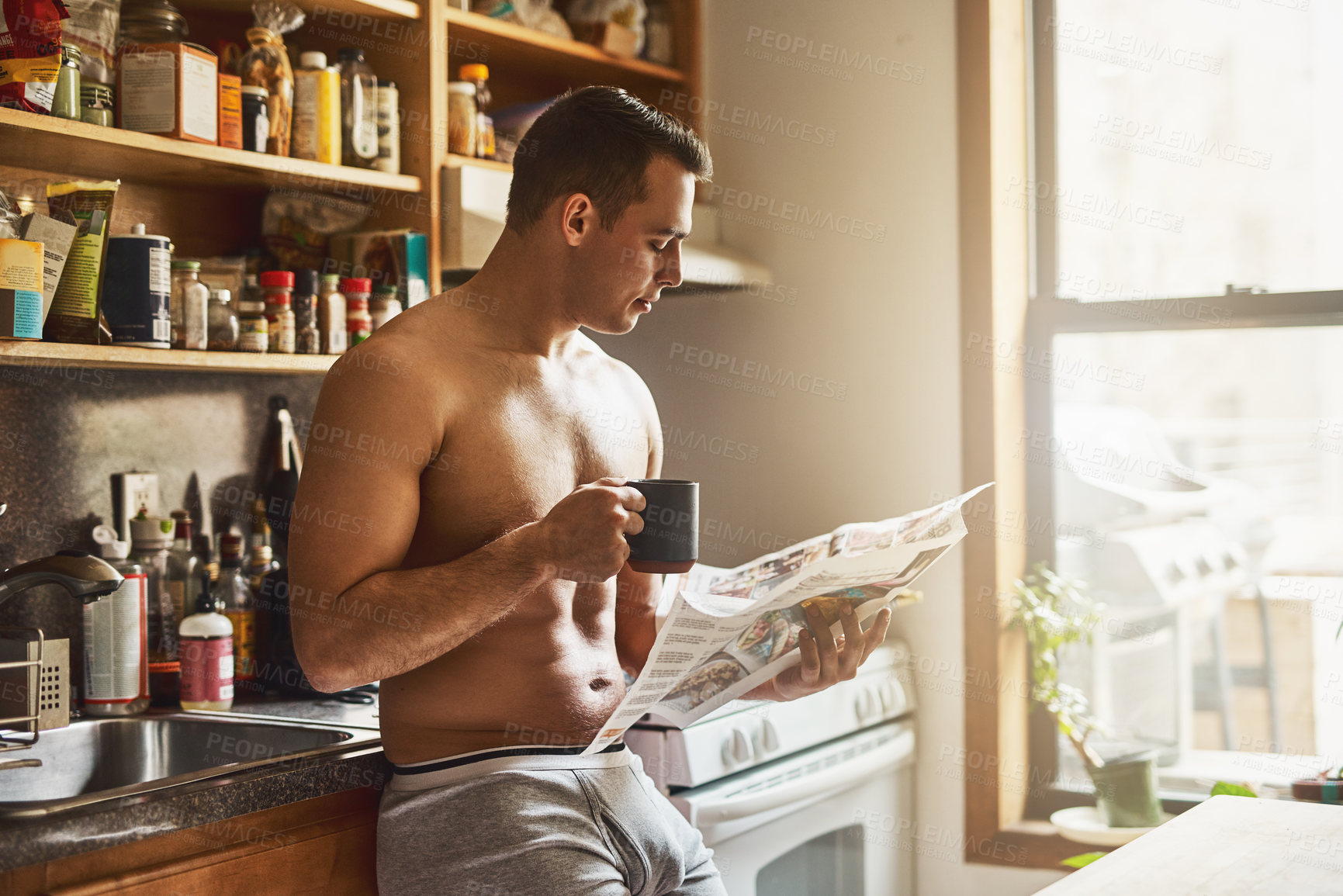 Buy stock photo Cropped shot of a handsome young shirtless man drinking a cup of coffee and reading the newspaper in the kitchen at home