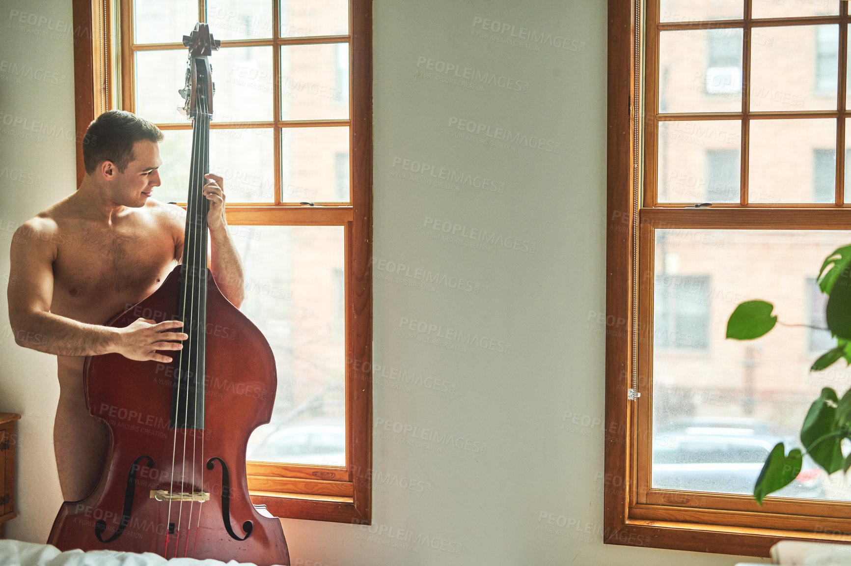 Buy stock photo Cropped shot of a young handsome shirtless man playing the cello and looking out the window at home
