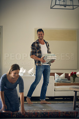 Buy stock photo Shot of a young beautiful woman rolling up a carpet while her handsome husband is carrying books at home