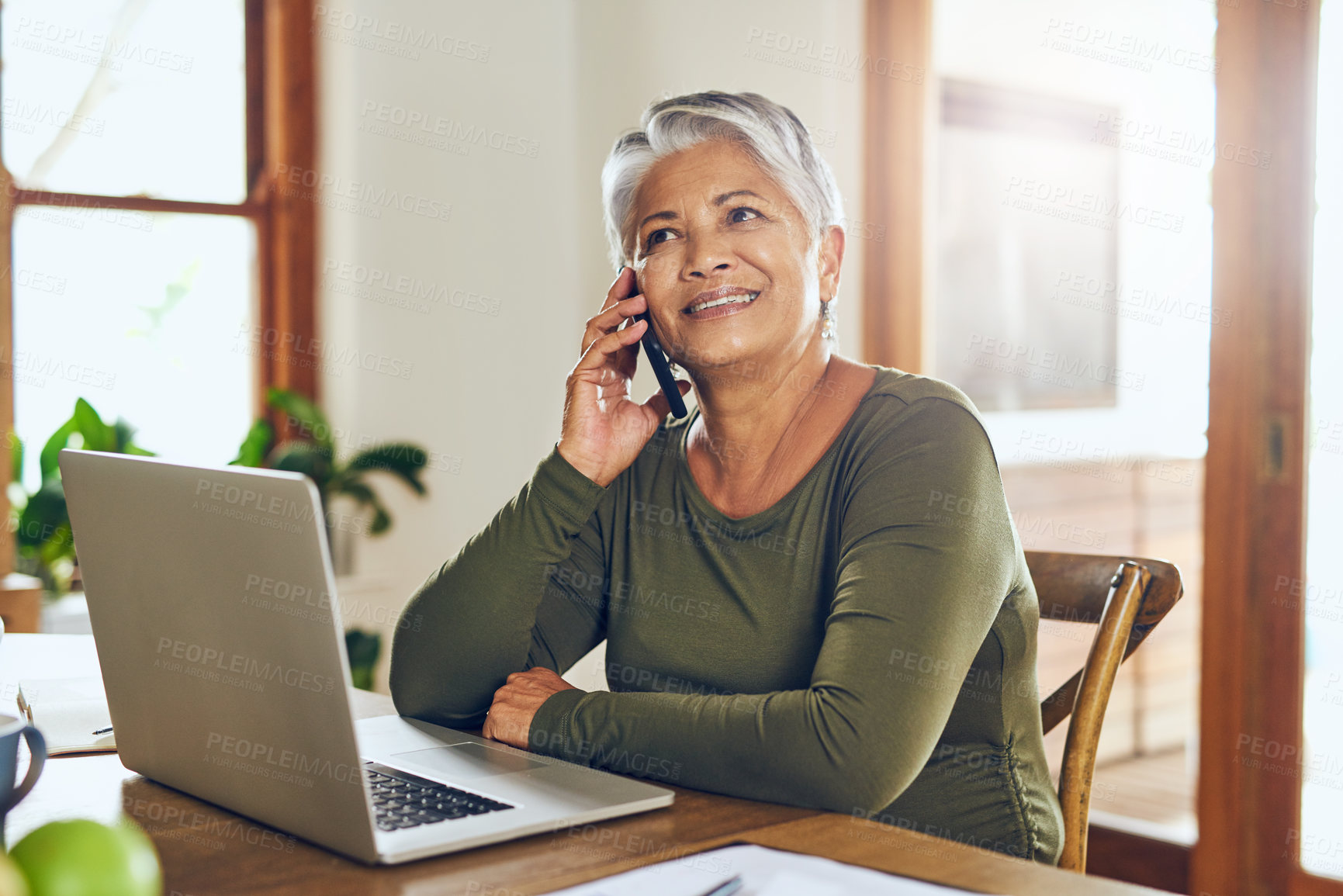 Buy stock photo Shot of a mature woman talking on a cellphone while working on a laptop at home