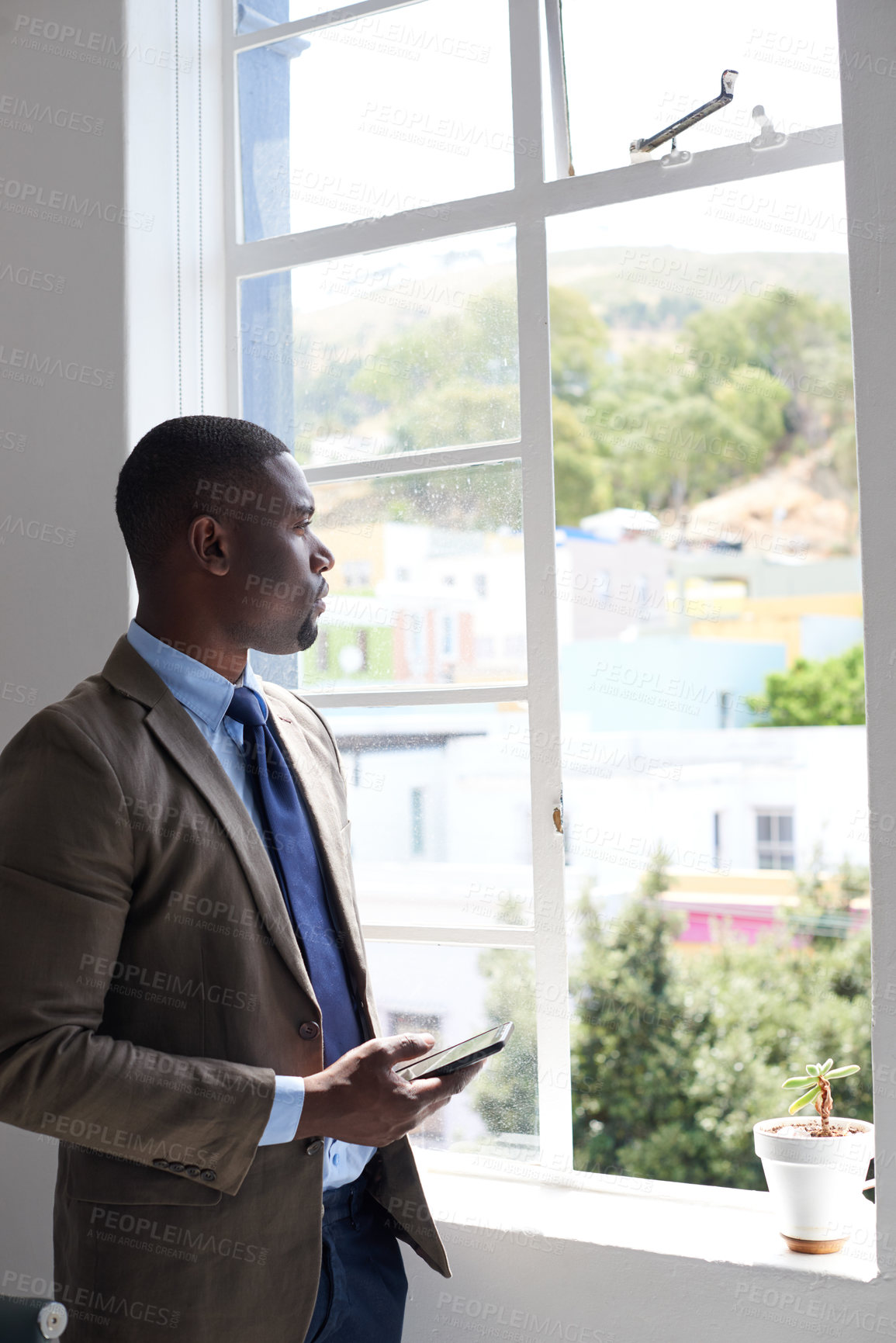 Buy stock photo Shot of a young businessman using a cellphone while looking out a window in an office