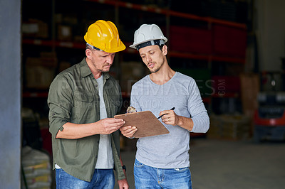 Buy stock photo Cropped shot of two male construction workers looking over some paperwork while standing in an industrial warehouse