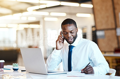 Buy stock photo Shot of a cheerful young businessman talking on his cellphone and making notes while being seated at his desk in the office during the day