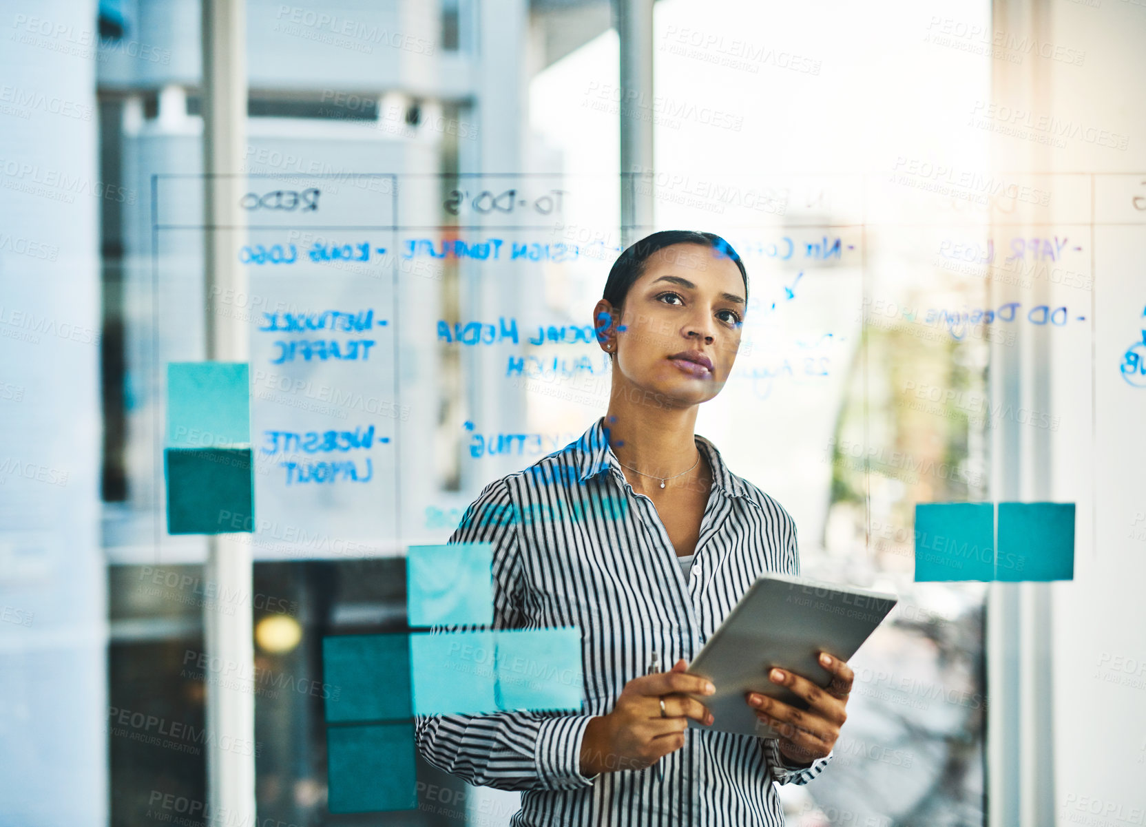 Buy stock photo Shot of a young businesswoman using a digital tablet while brainstorming on a glass wall in an office