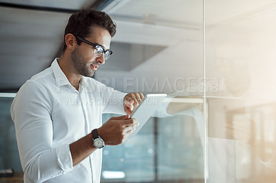 Buy stock photo Cropped shot of a handsome young businessman working on his digital tablet while standing in the office