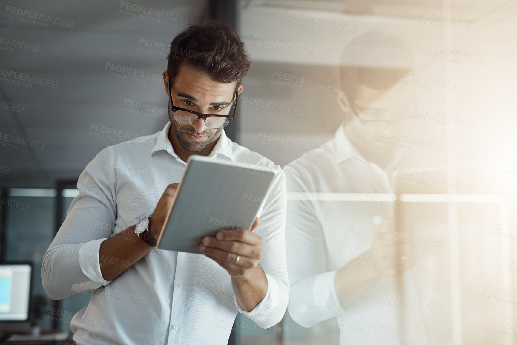 Buy stock photo Cropped shot of a handsome young businessman working on his digital tablet while standing in the office