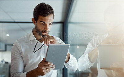 Buy stock photo Cropped shot of a handsome young businessman looking thoughtful while working on his digital tablet while standing in the office