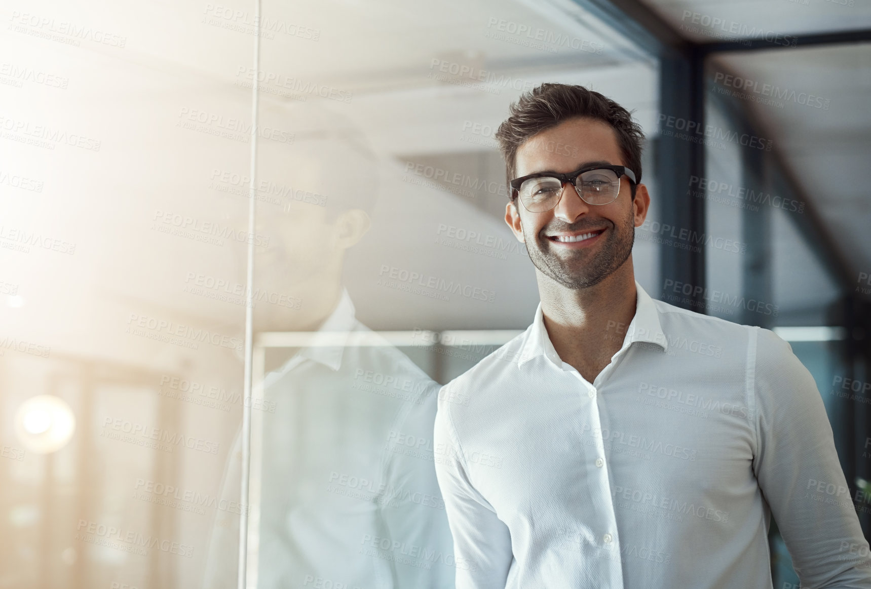 Buy stock photo Cropped portrait of a handsome young businessman leaning against a glass wall in his modern office