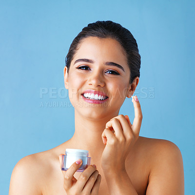 Buy stock photo Studio portrait of a beautiful young woman applying lotion to her face against a blue background
