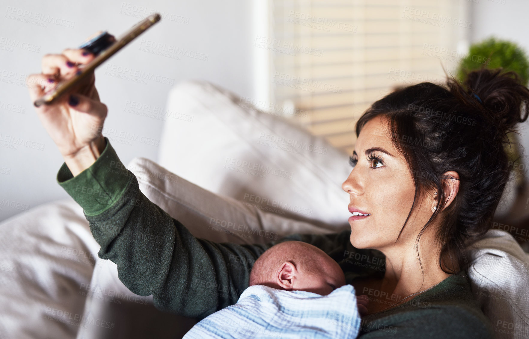 Buy stock photo Shot of a cheerful young woman lying on the sofa while holding her little infant son and taking a selfie at home during the day