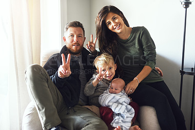 Buy stock photo Portrait of a cheerful young family seated on a sofa together while showing a peace sign at home during the day