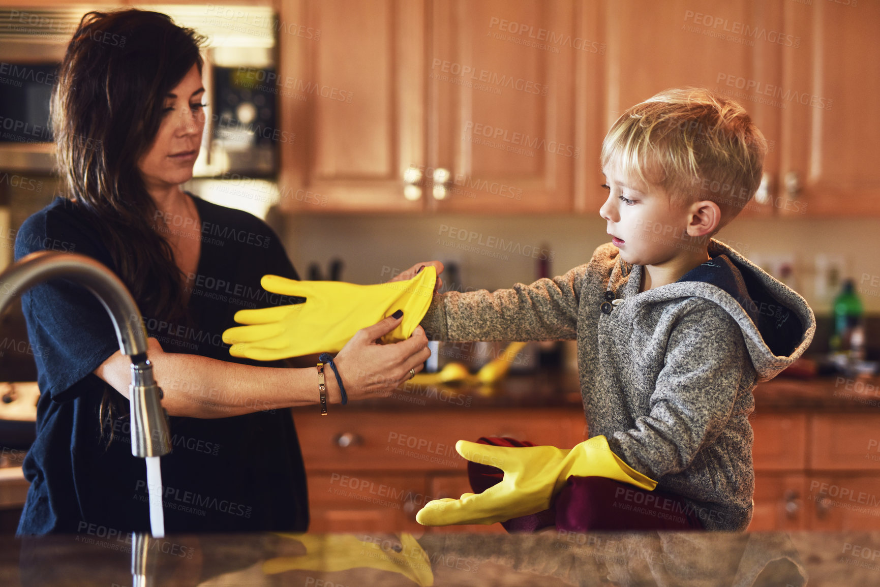 Buy stock photo Shot of a carefree young woman putting yellow washing gloves on her little boys hands to help her do the dishes at home during the day