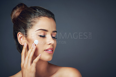 Buy stock photo Studio shot of a beautiful young woman applying moisturizer on to her face while posing against a blue background
