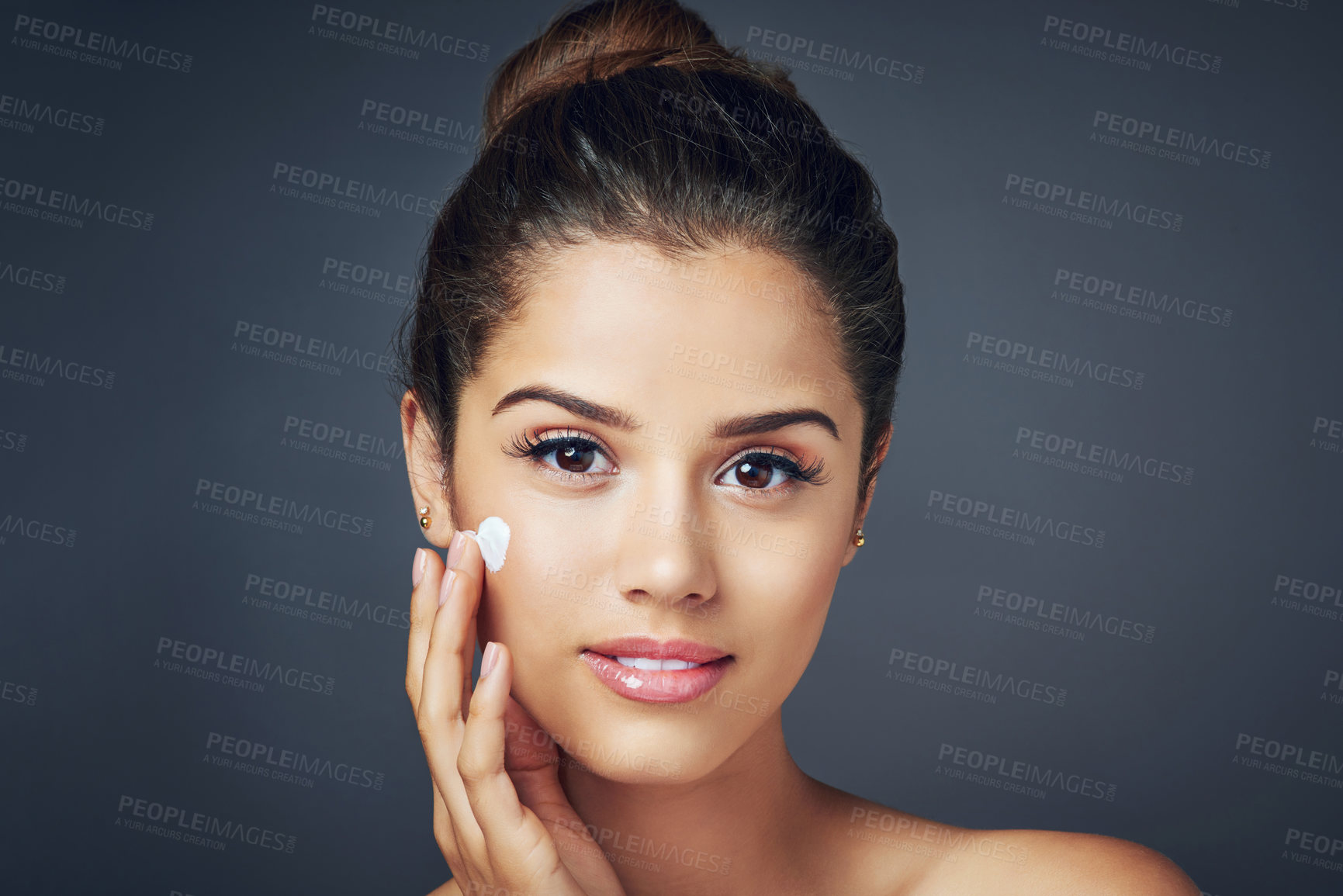 Buy stock photo Studio shot of a beautiful young woman applying moisturizer on to her face while posing against a blue background