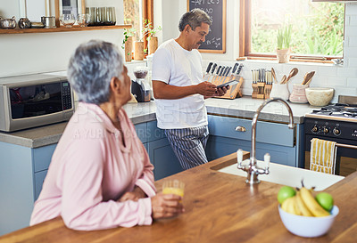 Buy stock photo Shot of an carefree elderly woman drinking orange jiuce in the kitchen while her husband is browsing on a digital tablet at home during the day