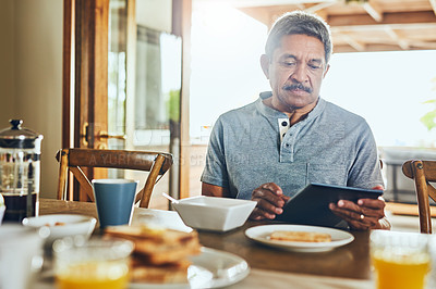 Buy stock photo Shot of a focused elderly man browsing on a tablet while having breakfast a table at home