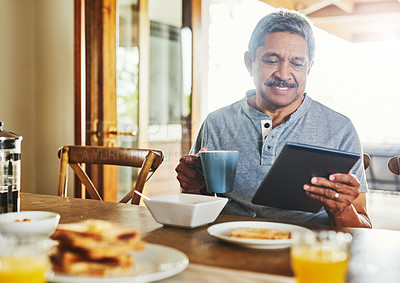 Buy stock photo Shot of a cheerful elderly man browsing on a tablet while having breakfast around  a table at home