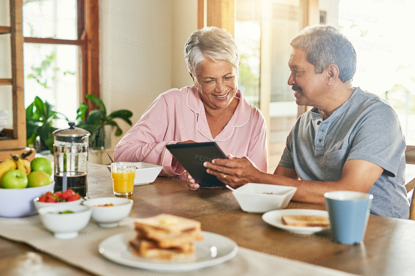 Buy stock photo Shot of a cheerful elderly couple browsing on a digital tablet together while having breakfast around a table at home