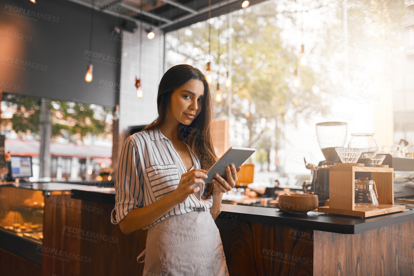 Buy stock photo Cropped portrait of an attractive young woman working on a digital tablet while standing in her coffee shop
