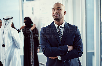 Buy stock photo Cropped shot of a handsome young businessman standing with his arms folded in the office with his colleagues in the background