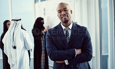 Buy stock photo Cropped portrait of a handsome young businessman standing with his arms folded in the office with his colleagues in the background
