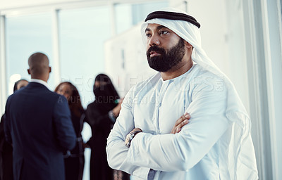Buy stock photo Cropped shot of a handsome mature arabic businessman standing with his arms folded in the office with his colleagues in the background