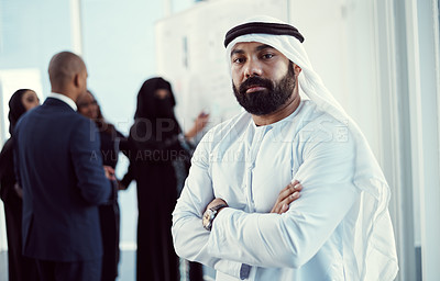 Buy stock photo Cropped portrait of a handsome mature arabic businessman standing with his arms folded in the office with his colleagues in the background