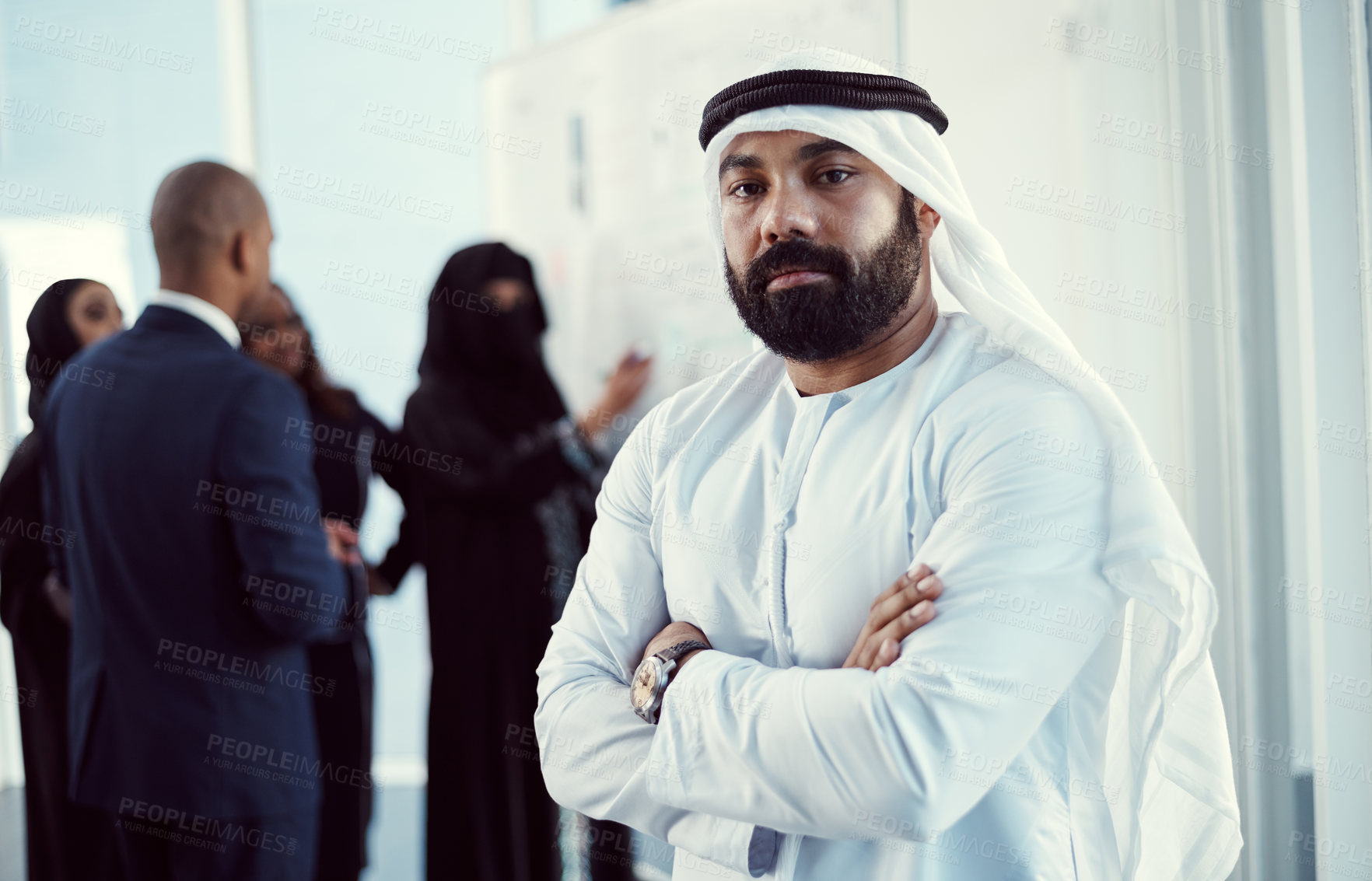 Buy stock photo Cropped portrait of a handsome mature arabic businessman standing with his arms folded in the office with his colleagues in the background