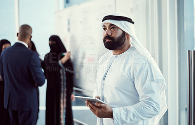 Buy stock photo Cropped shot of a handsome mature arabic businessman working on a tablet in the office with his colleagues in the background