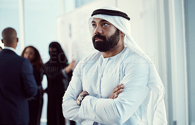 Buy stock photo Cropped shot of a handsome mature arabic businessman standing with his arms folded in the office with his colleagues in the background