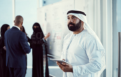 Buy stock photo Cropped shot of a handsome mature arabic businessman working on a tablet in the office with his colleagues in the background